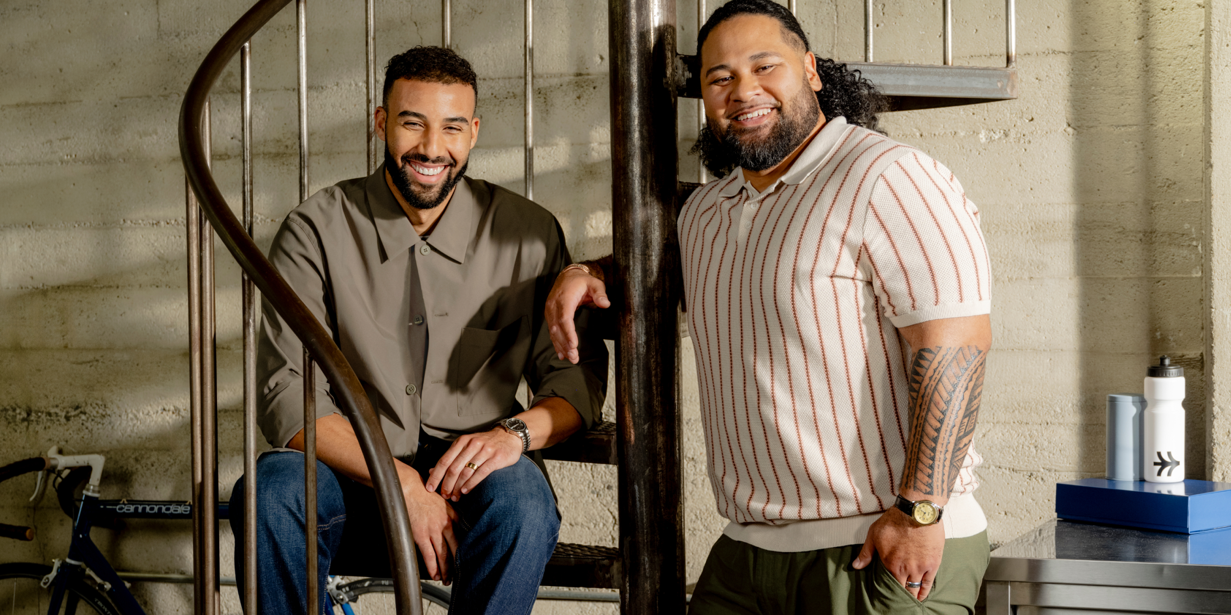 Two Shopify merchants smiling on a spiral staircase. One wears a gray shirt and jeans, while the other is in a striped polo and green pants. A bicycle is in the background, and a table holds a water bottle and a blue box.